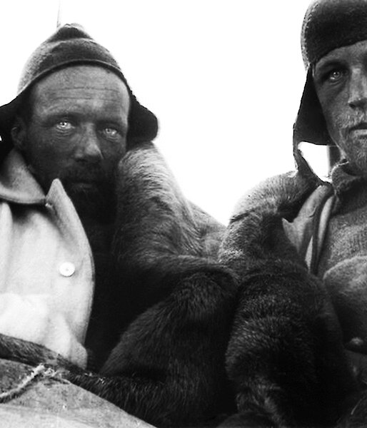 Two men wearing woollen beanies and hunching to keep warm stare at the camera with weathered faces.