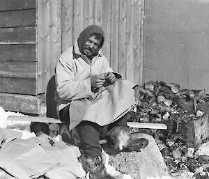 Frank Hurley wearing furry boots and warm hood smiles at the camera while he sits outside a hut in the sun sewing.