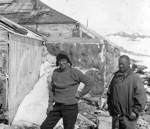 Two expeditioners standing outside a snow-covered wooden hut