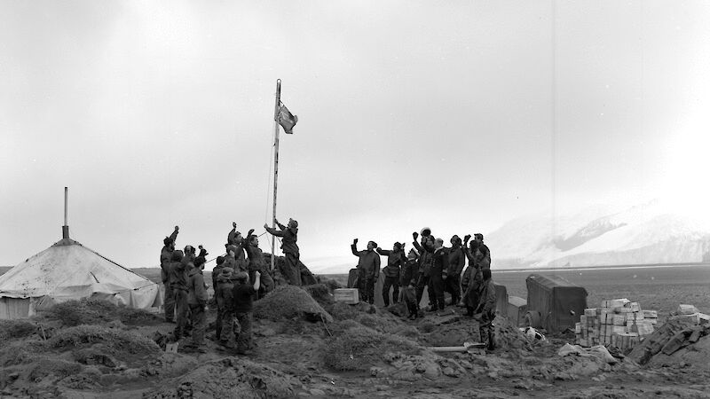 Black and white image of expedition leader raising flag surrounded by cheering men