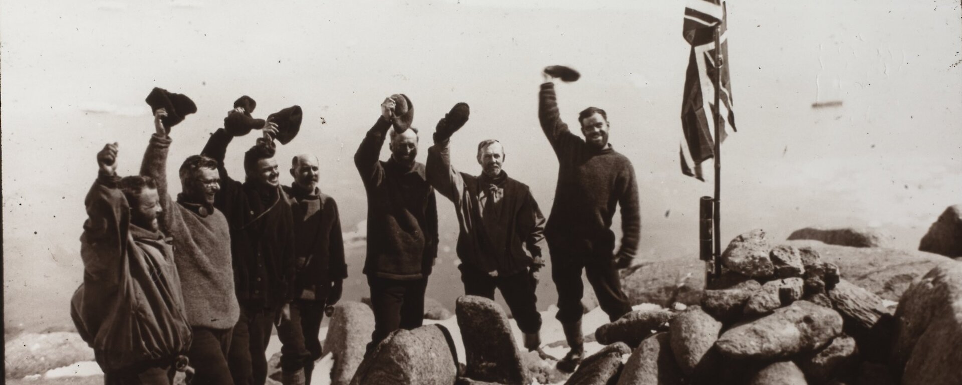 A black and white photograph of group standing next to a flag and waving their hats.