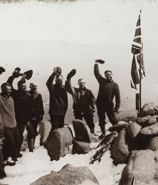 A black and white photograph of group standing next to a flag and waving their hats.