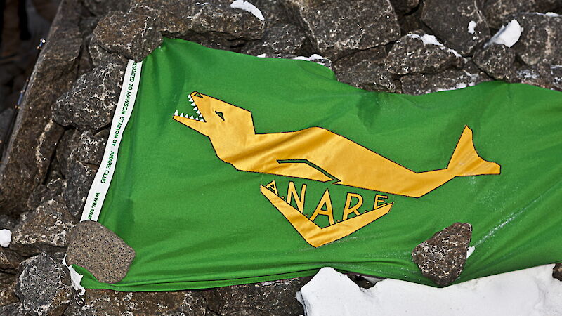 A triangle shaped flag with seal and letters ANARE surrounded by rocks