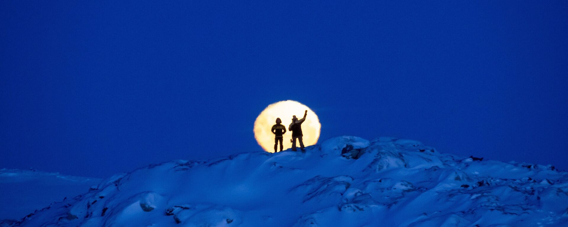 Two expeditioners silhouetted against a bright yellow moon standing atop a snowy mountain. Yellow moon stands out against a royal blue sky and mountain.