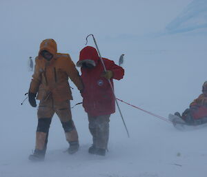 Two scientists walking in mist with shepherds’ crooks, pulling other expeditioner on sled