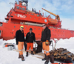 Expeditioners posing beside stack of wood and kindling and BBQ on the ice beside the ship.