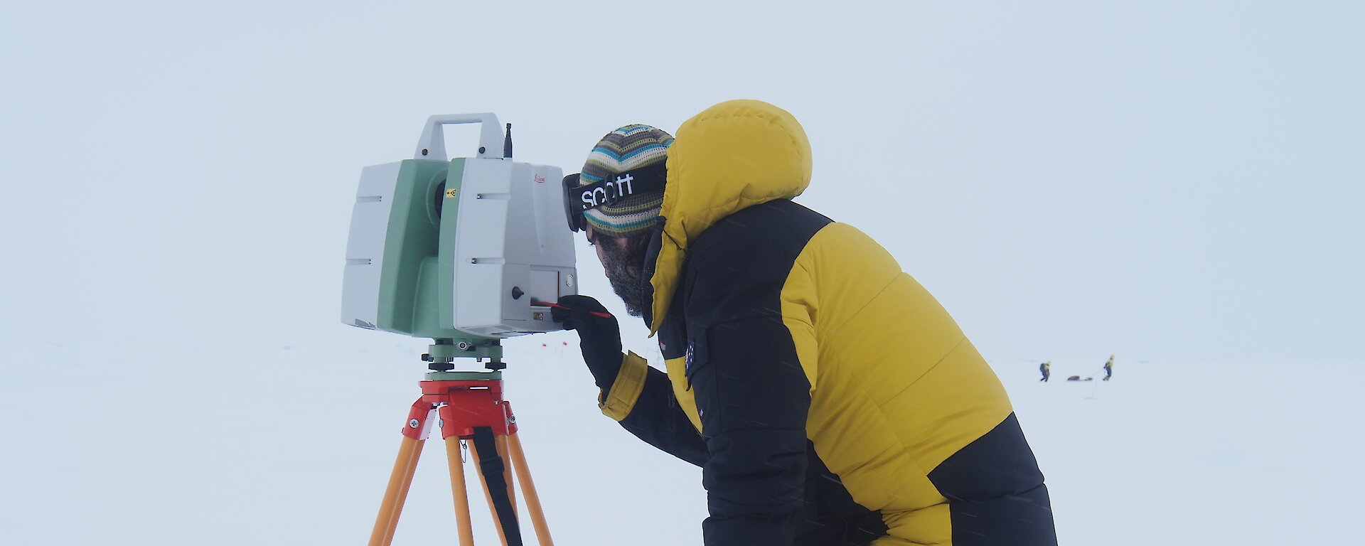 A scientist wearing cold weather gear looks into a scanner on top of a tripod on the ice.