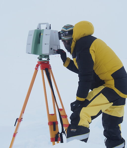 A scientist wearing cold weather gear looks into a scanner on top of a tripod on the ice.