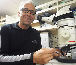 Scientists smiles at the camera while sitting at the microscope.