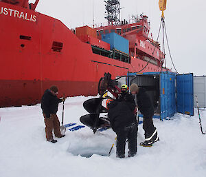 The ship stands by as the crew on the ice stand around to dig the hole in the ice.