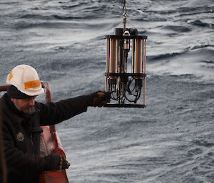 The bosun pushes a large metal device strung from a hook over the water.