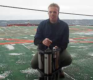 A young man kneeling on the deck of the ship holding a large metal device in front of him.