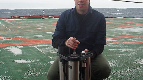 A young man kneeling on the deck of the ship holding a large metal device in front of him.