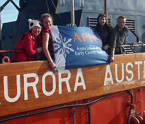 Four young scientists are holding up a flag with saying Association of early career scientists above the rail and the sign of the Aurora Australis