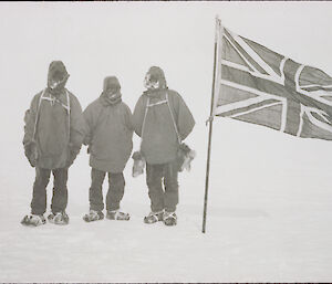 Black and white image of three men in polar clothing posed with British flag at their farthest south position on the ice cap