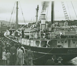 Black and white image of small wooden ship arriving at pier with crowd surrounding