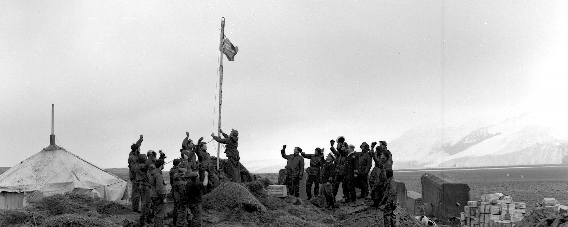 Black and white image of expedition leader raising flag surrounded by cheering men