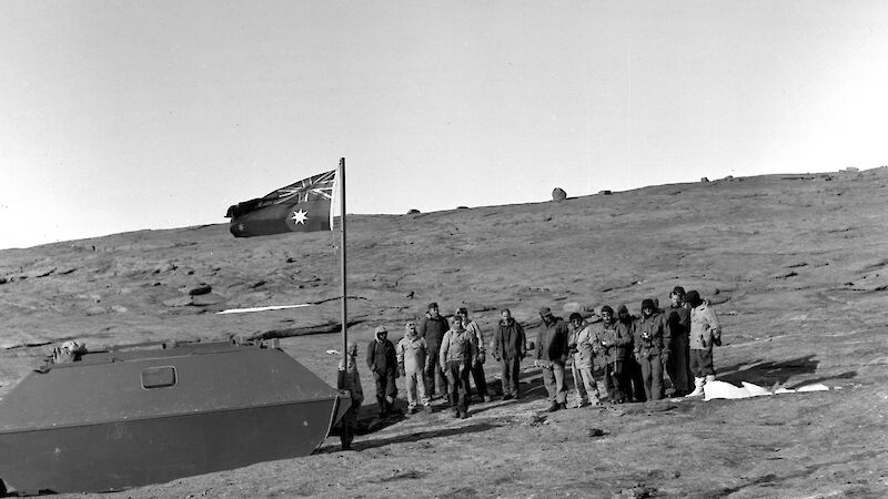 Black and white image from 1954 showing a group of men next to a flag pole with old Australian flag and low structure