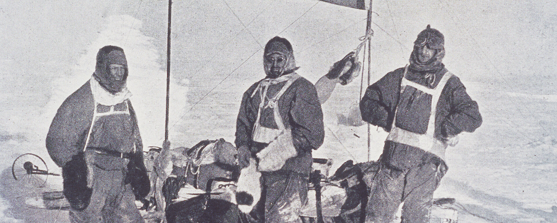 Black and white photo of three men in old-fashioned polar clothes by a sledge.