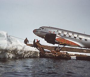 A silver plane is unloaded from a boat onto the icy shore.