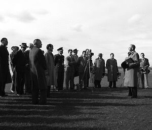 Vintage photo of a varied group of men, some in trench coats with large cameras, listen to a single male speaker