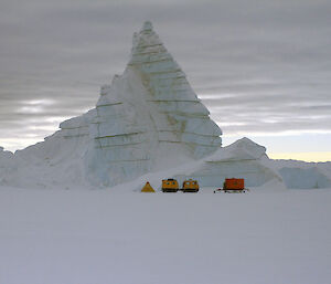 Orange and yellow vehicles below a towering ice formation.