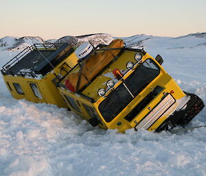 A yellow oversnow vehicle tilted to one side, stuck in a ditch