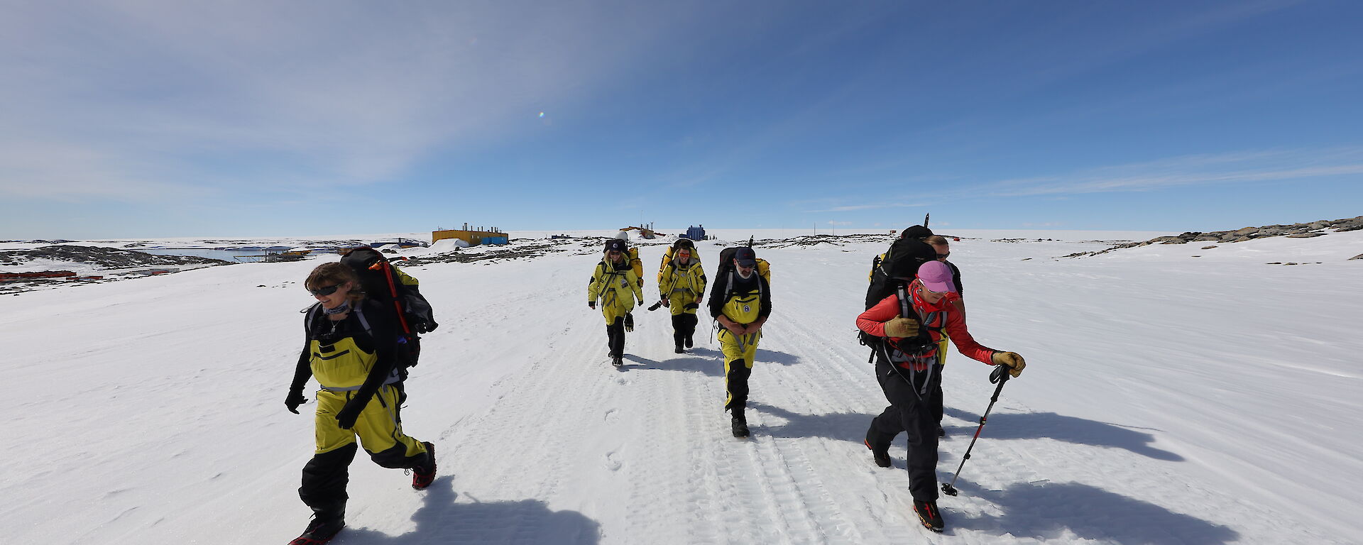 A group of expeditioners walk through an icy landscape under a clear, blue sky.