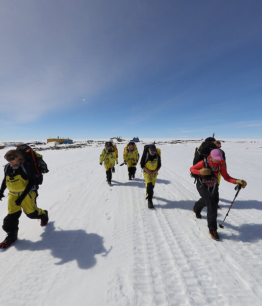 A group of expeditioners walk through an icy landscape under a clear, blue sky.