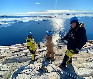 3 expeditioners stand on rocky ground overlooking a blue ocean with frozen patches.