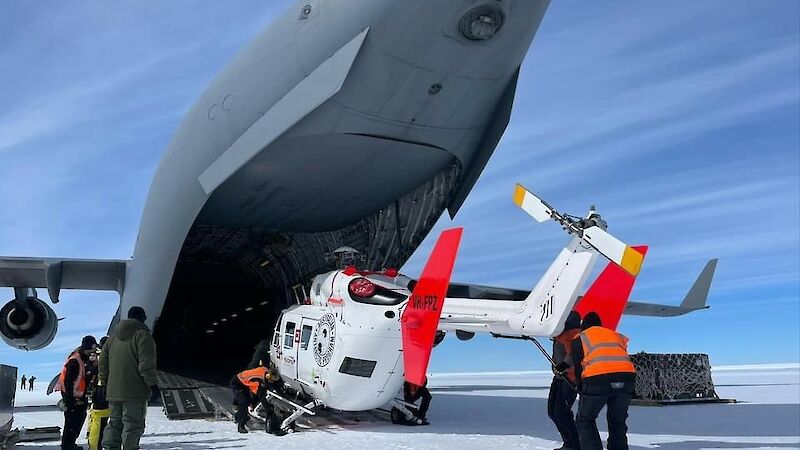 A helicopter is rolled out of the back of a large grey air force plane