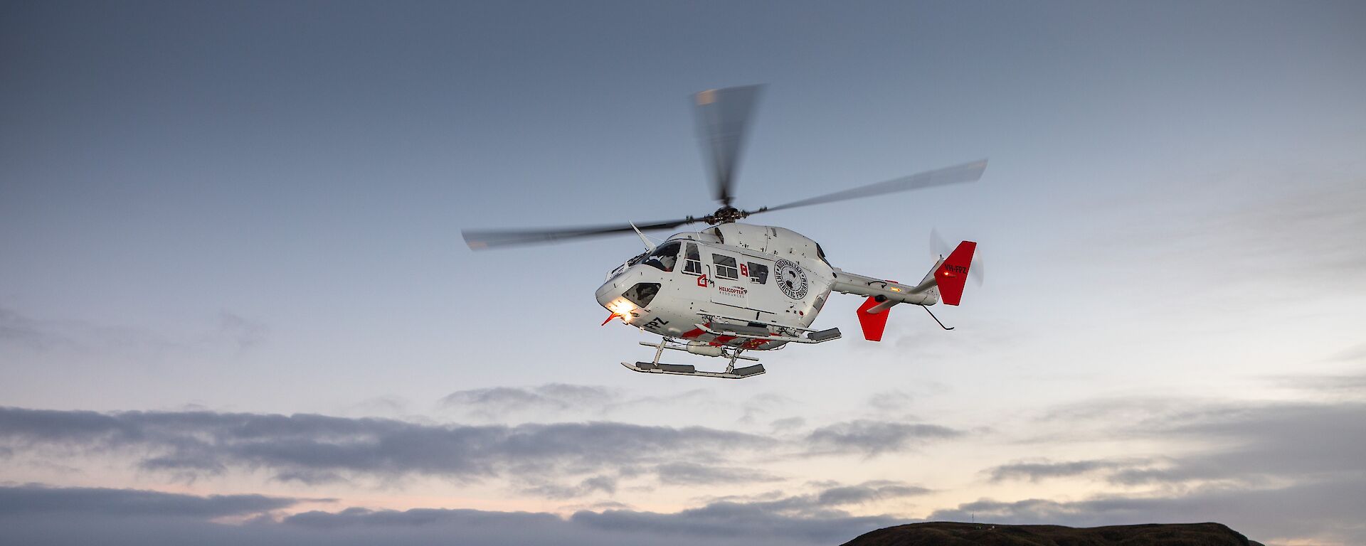 A white helicopter takes off from helipad at dusk with an island behind it