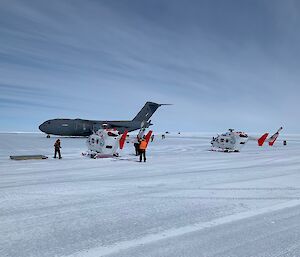 Two white helicopters with red tails sit on ice with a huge grey airforce plane behind them