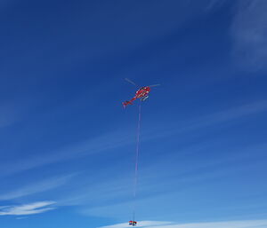 Red helicopter with slingload backed by a deep blue sky above the flat expanse of Totten Glacier