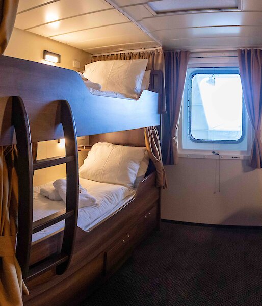 A small cabin room with bunk bed, a cupboard, a window and a desk with shelves above.