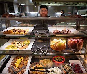 A selection of food displayed in refrigerated shelves. A man with a chef's cap stands behind in the kitchen.