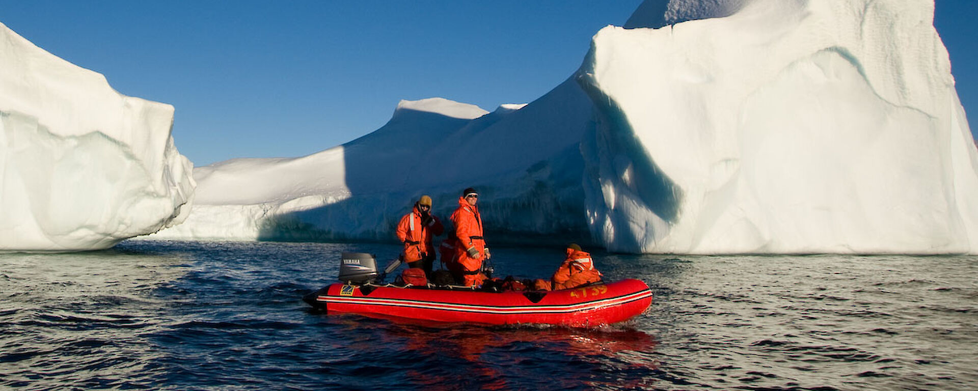 Two people in a red inflatable boat are dwarfed by a huge red iceberg.