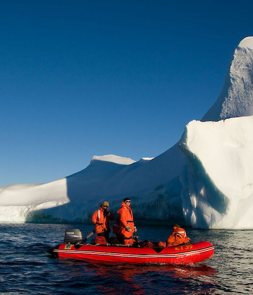 Two people in a red inflatable boat are dwarfed by a huge red iceberg.