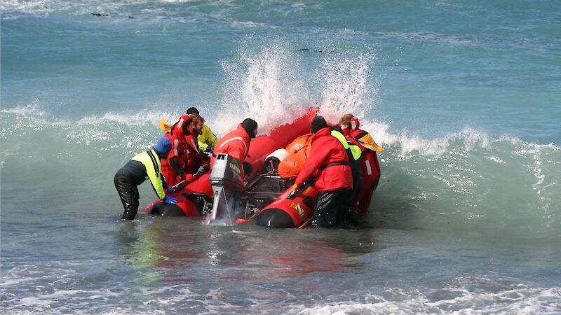 About six expeditioners in wet suits and gear wade in the water to push an inflatable boat over a wave to launch