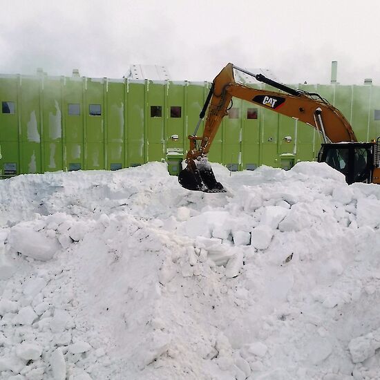 An excavator clears snow in foreground with a large green building in background