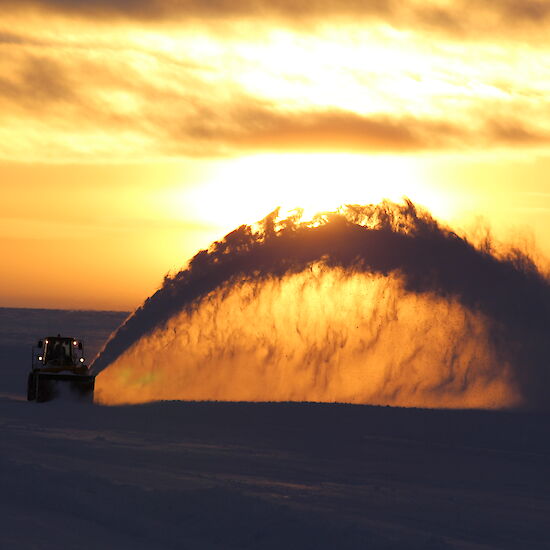 Machinery clearing runway at dusk with snow being projected out like a fountain.
