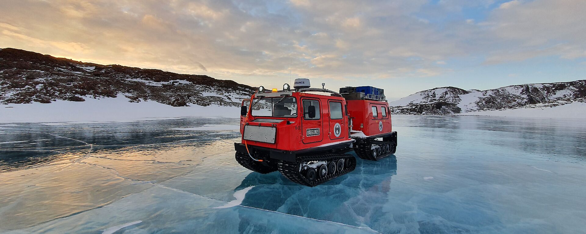 A red Hägglunds sits on the shiny blue frozen ice of a river with snow covered hills in the background