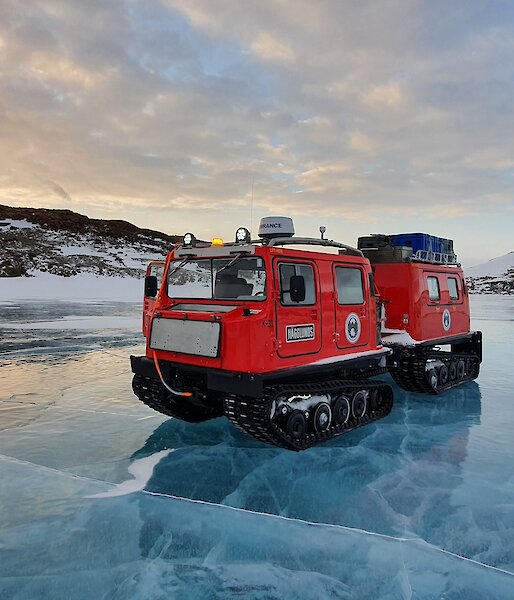 A red Hägglunds sits on the shiny blue frozen ice of a river with snow covered hills in the background