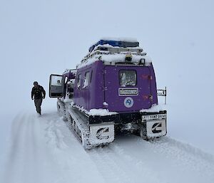 A purple vehicle is stopped on the snow. An expeditioner walks towards the open vehicle door.
