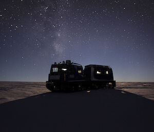 A Hägglunds vehicle is parked on the ice casting a large shadow. The sky is full of stars behind it.