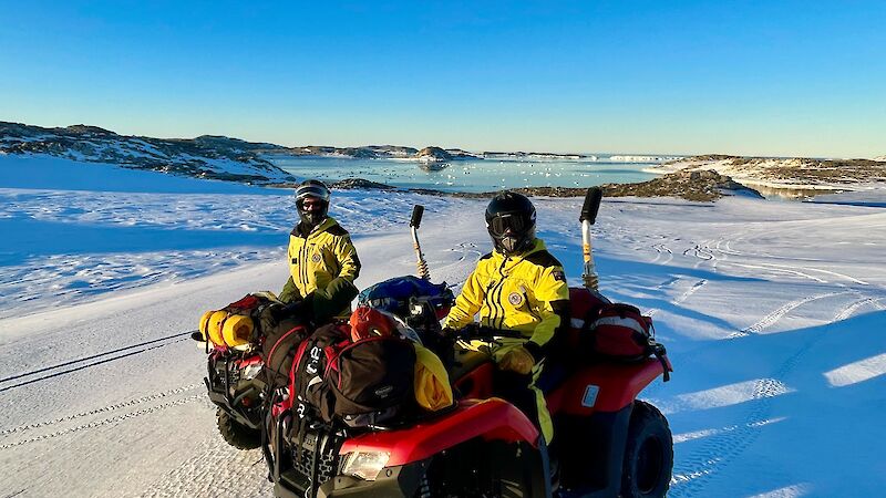2 expeditioners riding quads in an icy landscape on a sunny day. The quads are laden with gear.