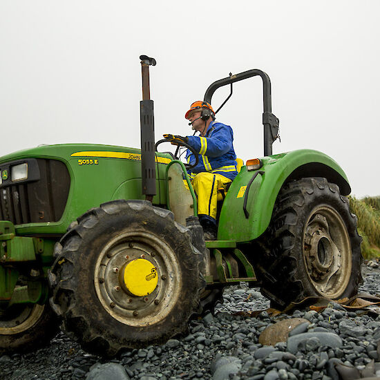 Expeditioner in blue jacket and orange hard hat driving green tractor on rocky beach whilst another expeditioner in black and orange suit and yellow helmet looks on