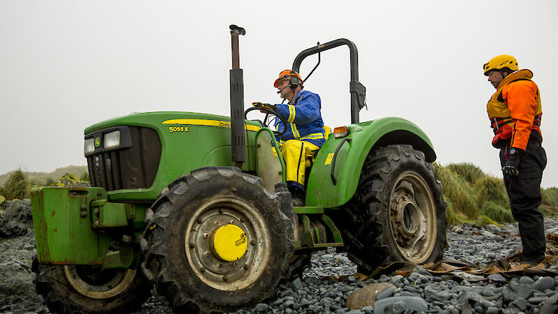 Expeditioner in blue jacket and orange hard hat driving green tractor on rocky beach whilst another expeditioner in black and orange suit and yellow helmet looks on