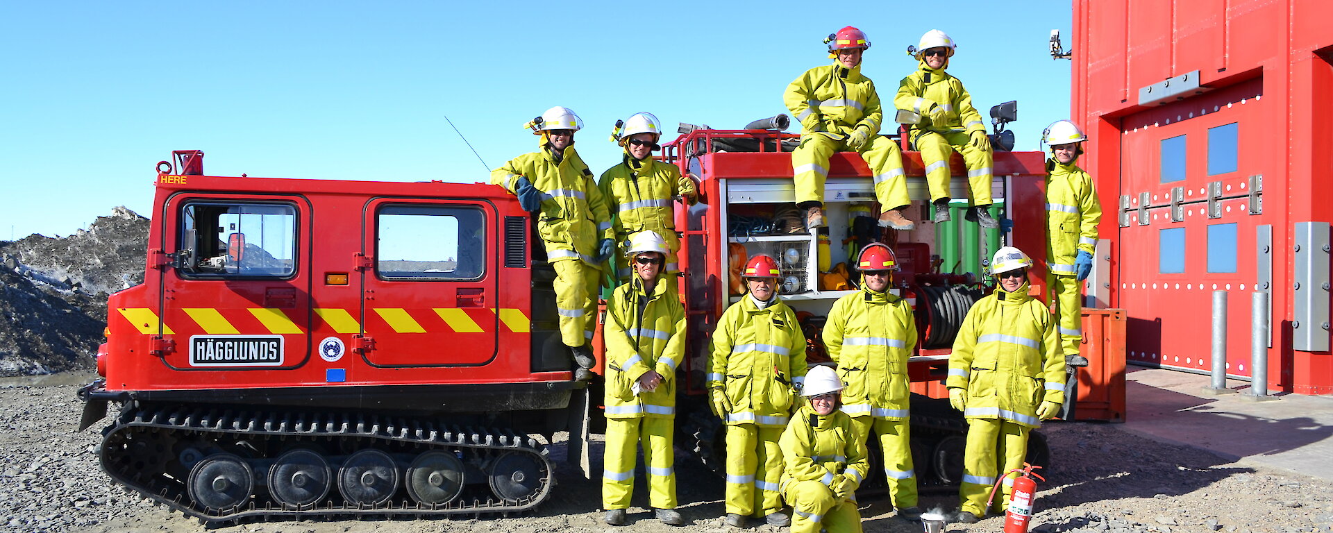 Expeditioners in fire fighting gear pose near and on the fire Hägglunds.