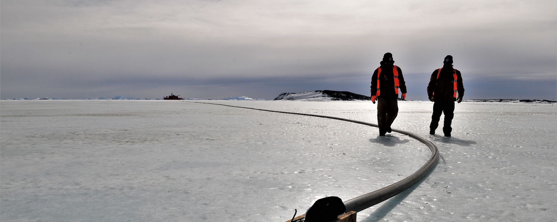 Two expeditioners walking along the fuel line with ship in background